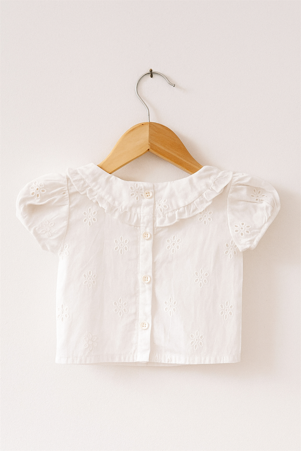 A white short-sleeved toddler blouse with delicate floral eyelet embroidery, buttoned down the back, is displayed on a wooden hanger against a light-colored wall. The top features a ruffled Peter Pan collar and puffed sleeves, giving it a soft, vintage-inspired charm.