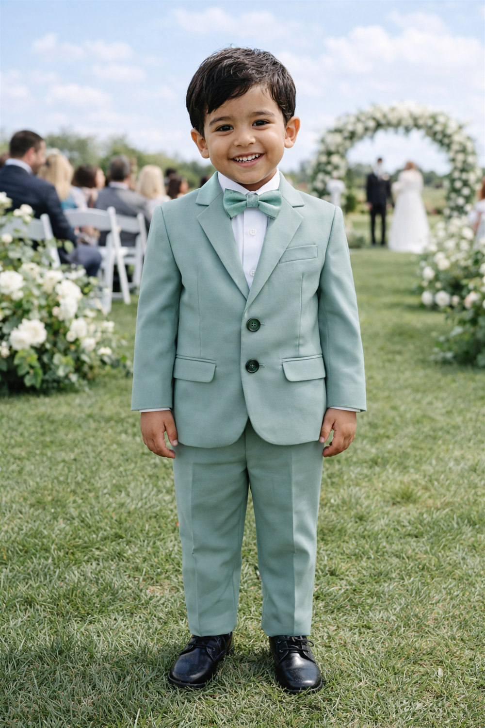 Smiling toddler boy in mint green suit and bow tie standing outdoors at wedding aisle with floral arches and guests.