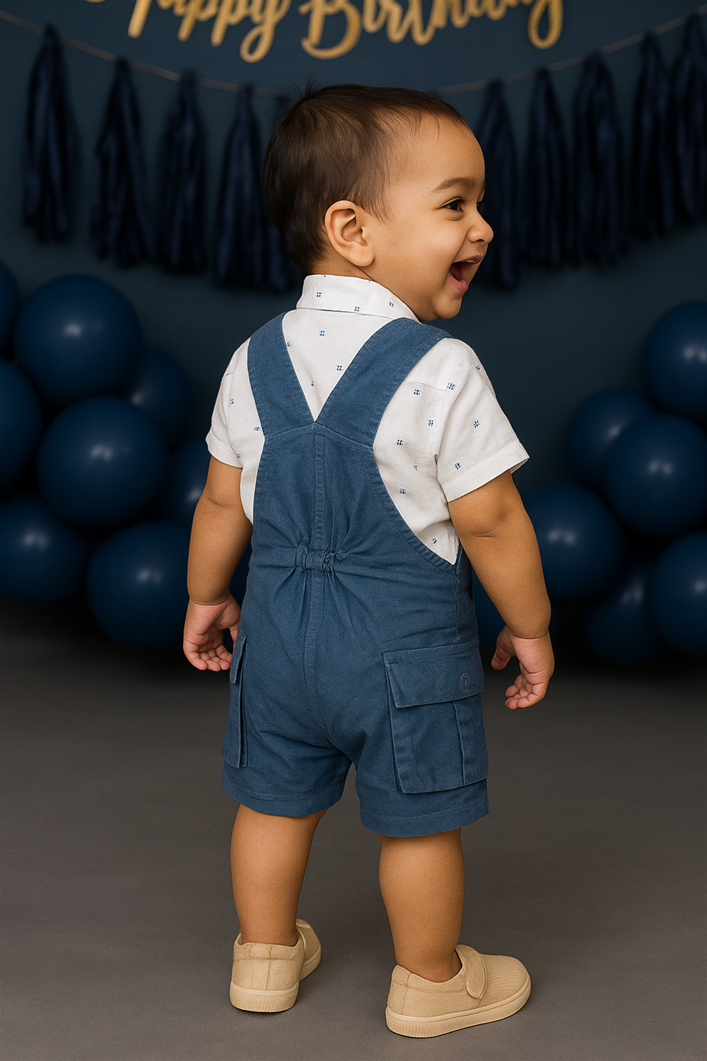 Back side view of a baby boy Wearing steel blue corduroy dungaree and a white shirt, sitting comfortably with a playful expression.