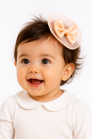 Smiling baby girl wearing peach bow hair clip on side of head, dressed in white outfit, looking adorable and happy.