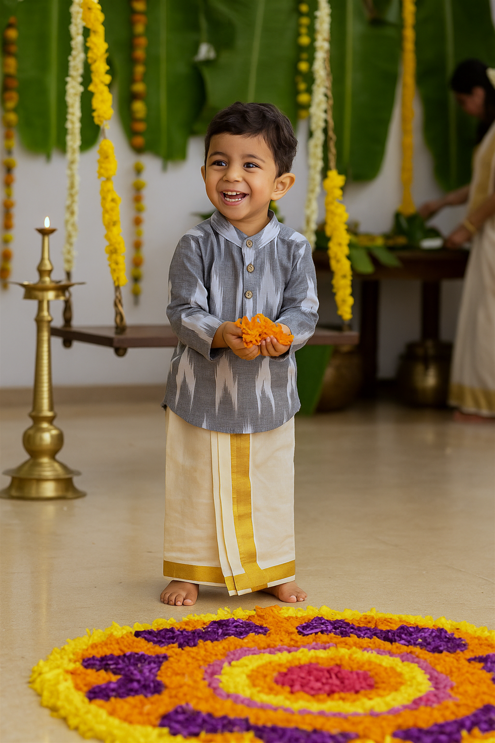 Smiling boy wearing grey ikkat kurta and cream dhoti, holding orange flowers, standing beside bright Onam floral pookalam.