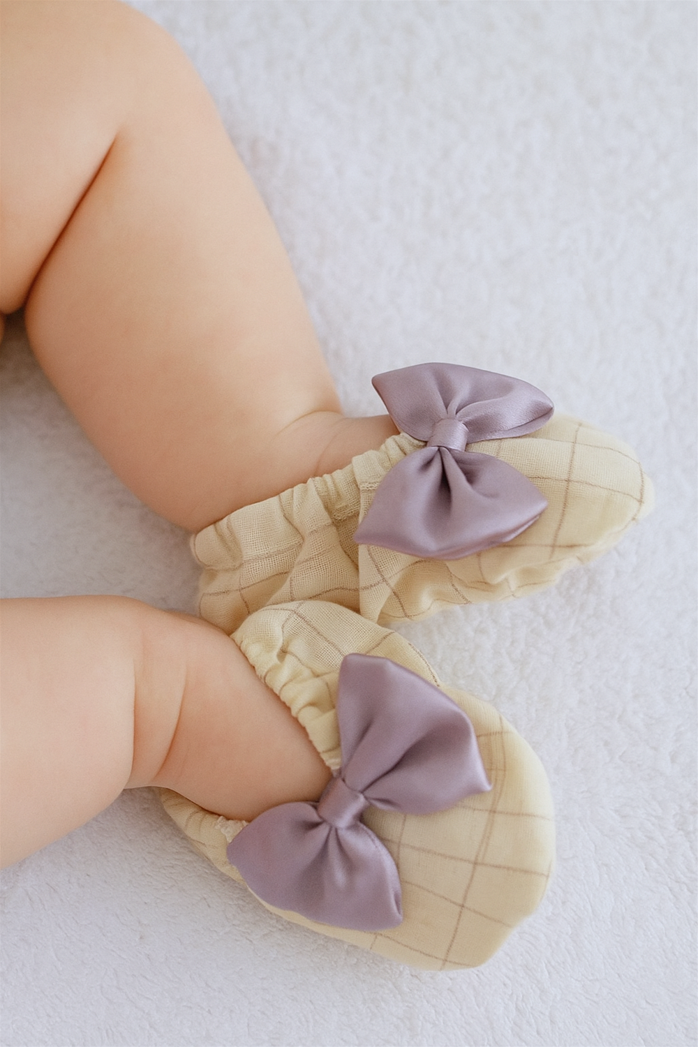 The baby booties are shown on a baby lying down, with chubby legs complementing the cozy cream shoes. The lavender bows create a stylish contrast against the light checkered fabric.