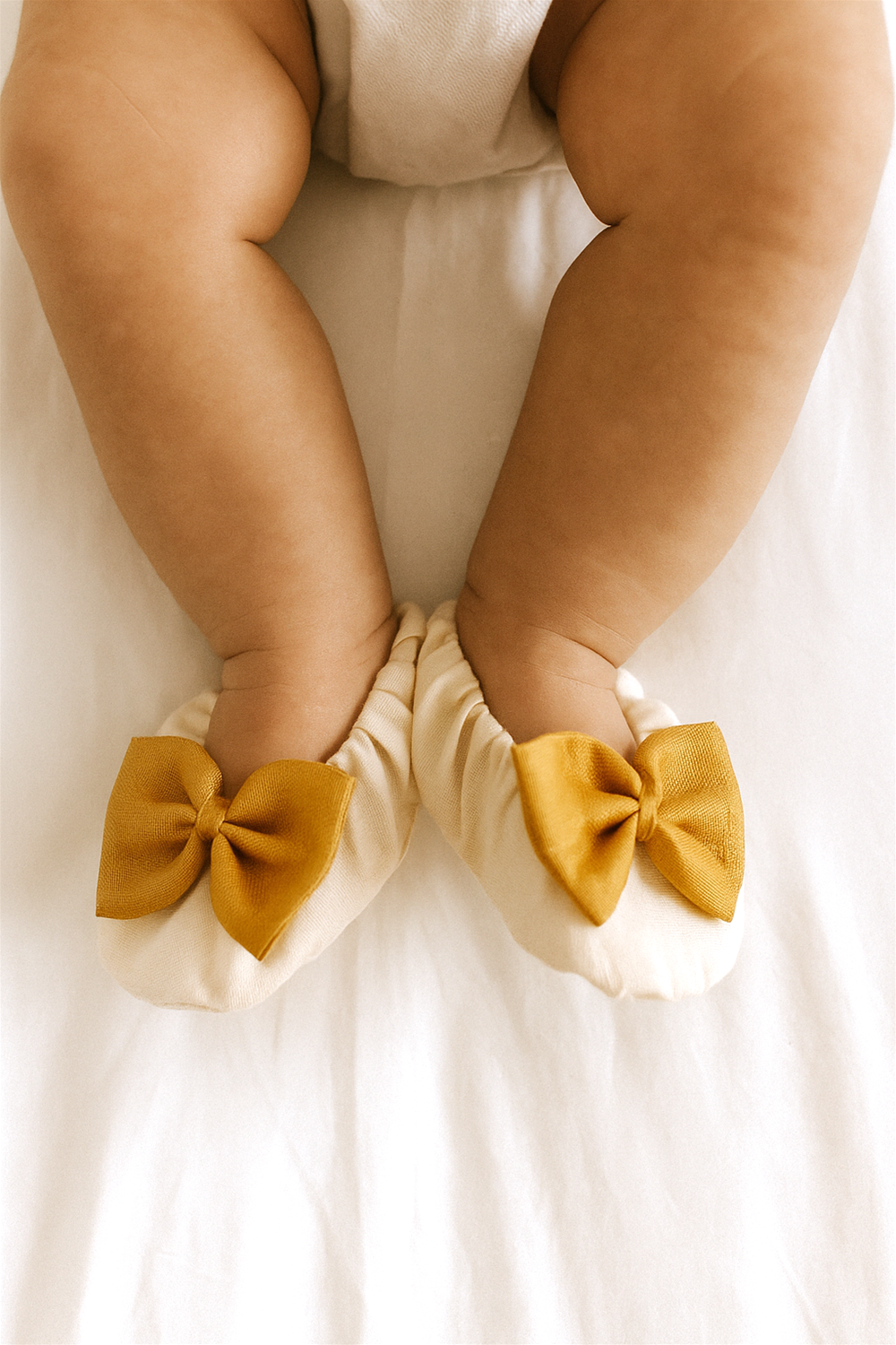A baby is wearing the cream booties with golden bows while lying on a bed. The warm lighting emphasizes the cozy and elegant look of the shoes on tiny feet.