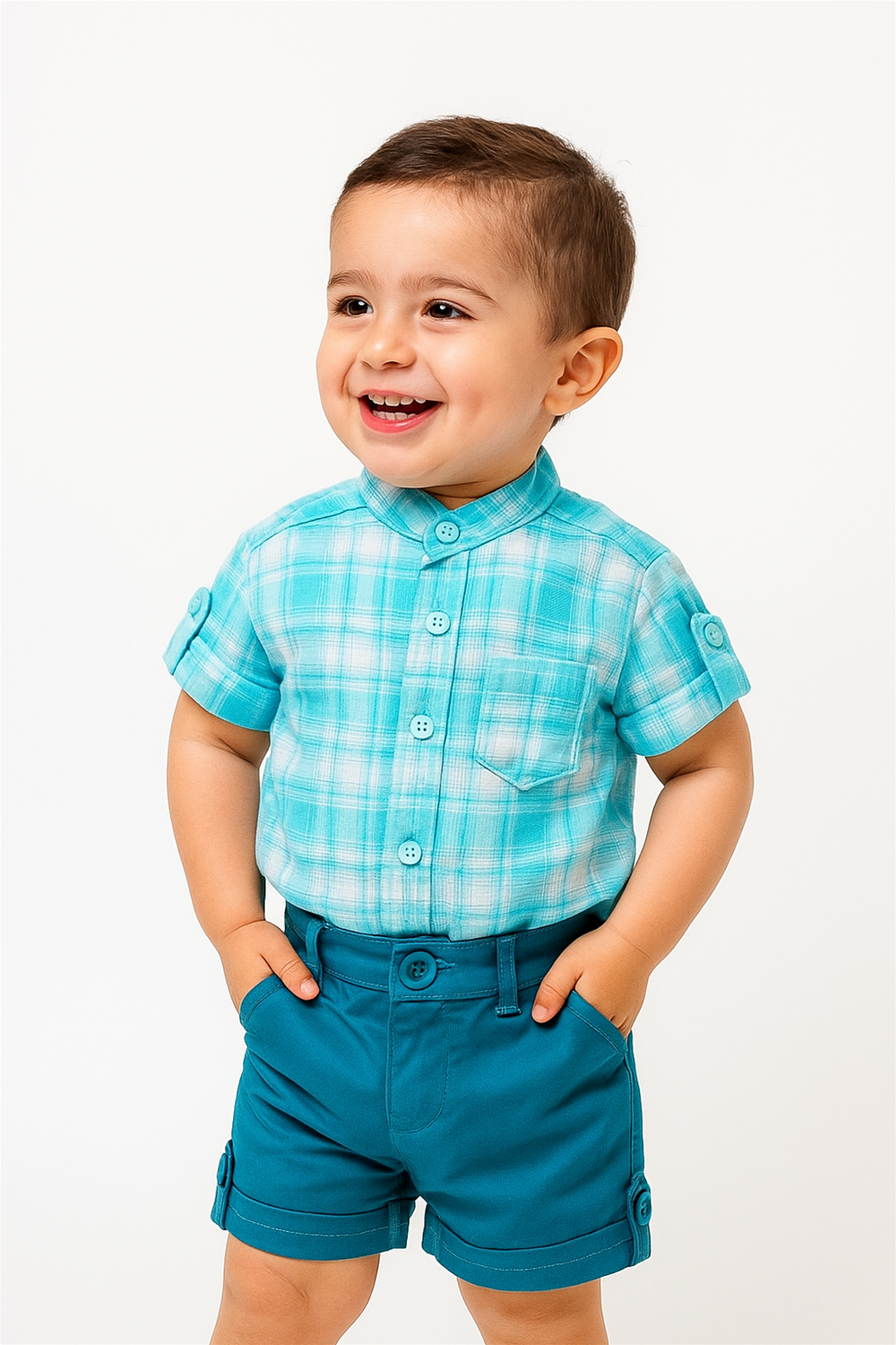 Smiling toddler boy wearing aqua plaid shirt and teal shorts, hands in pockets, standing against bright white studio background.
