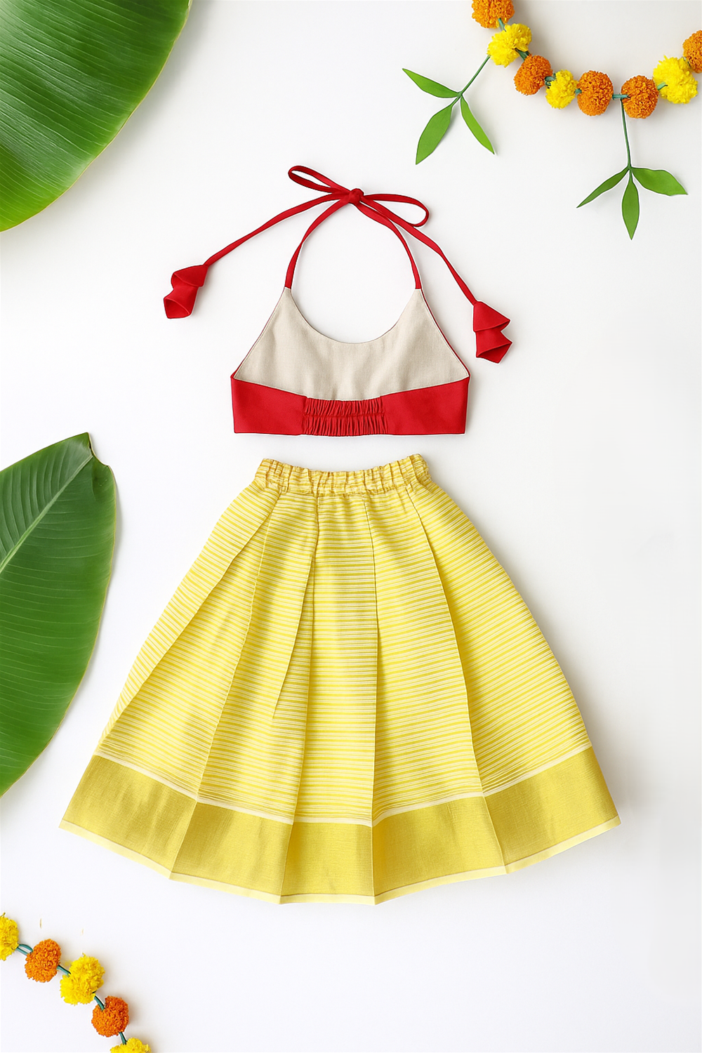 A traditional baby outfit is laid flat, featuring a red and beige halter-neck blouse with tassels and a yellow striped skirt. Decorative elements like banana leaves, marigold garlands, and brass lamps frame the festive presentation.
