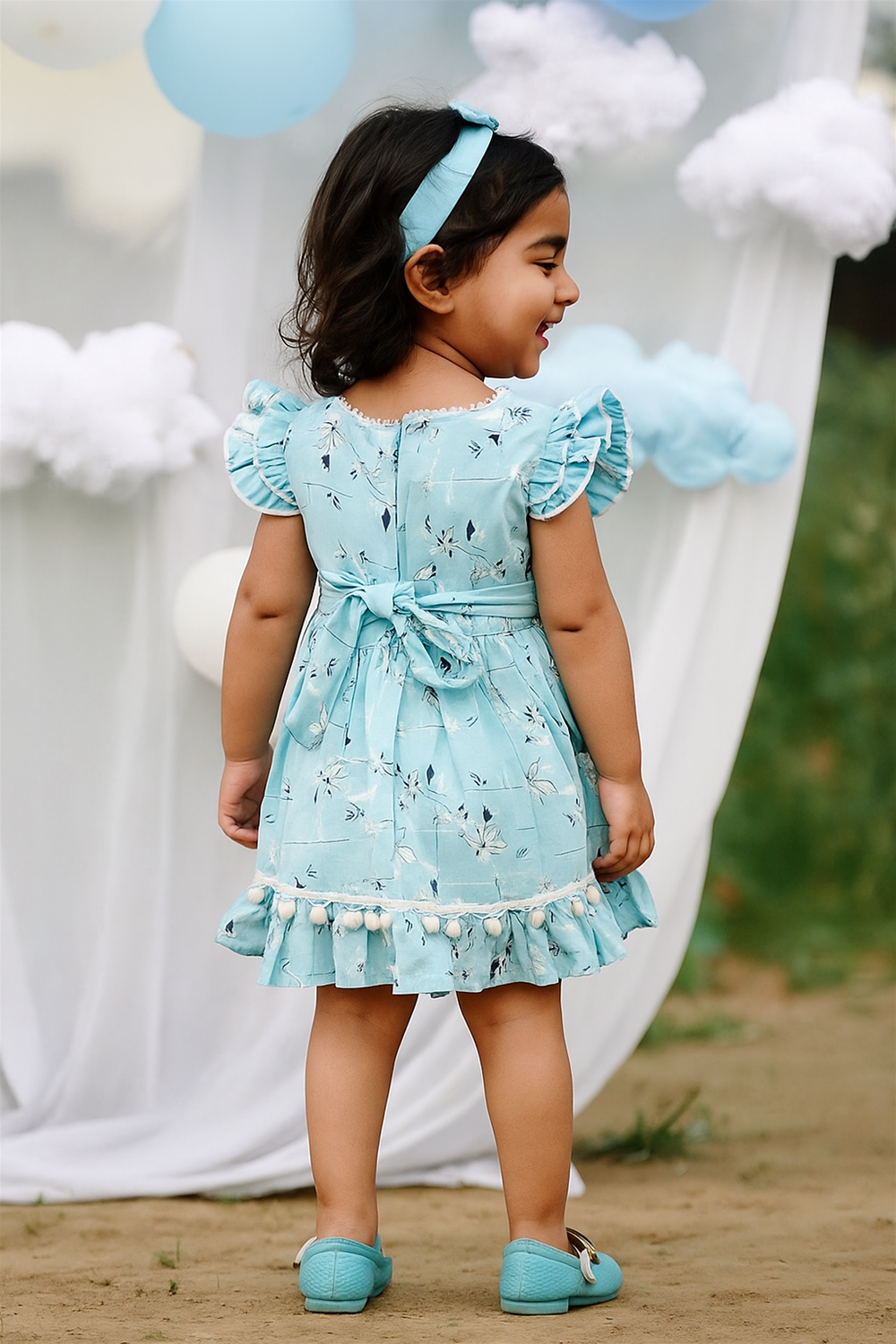 A young girl facing away, wearing a sky blue floral frock with flutter sleeves, pom-pom trim, and a back bow. She stands smiling in front of a dreamy setup with clouds and balloons.