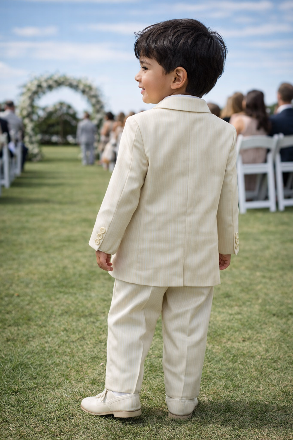 Toddler boy wearing formal suit walking outdoors on grass, seen from behind at a garden wedding ceremony setup.