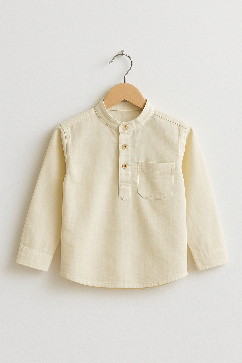A cream-colored boy’s kurta with a Chinese collar and wooden buttons is displayed on a wooden hanger. The kurta features a front chest pocket and long sleeves, set against a plain white background.A cream-colored boy’s kurta with a Chinese collar and wooden buttons is displayed on a wooden hanger. The kurta features a front chest pocket and long sleeves, set against a plain white background.
