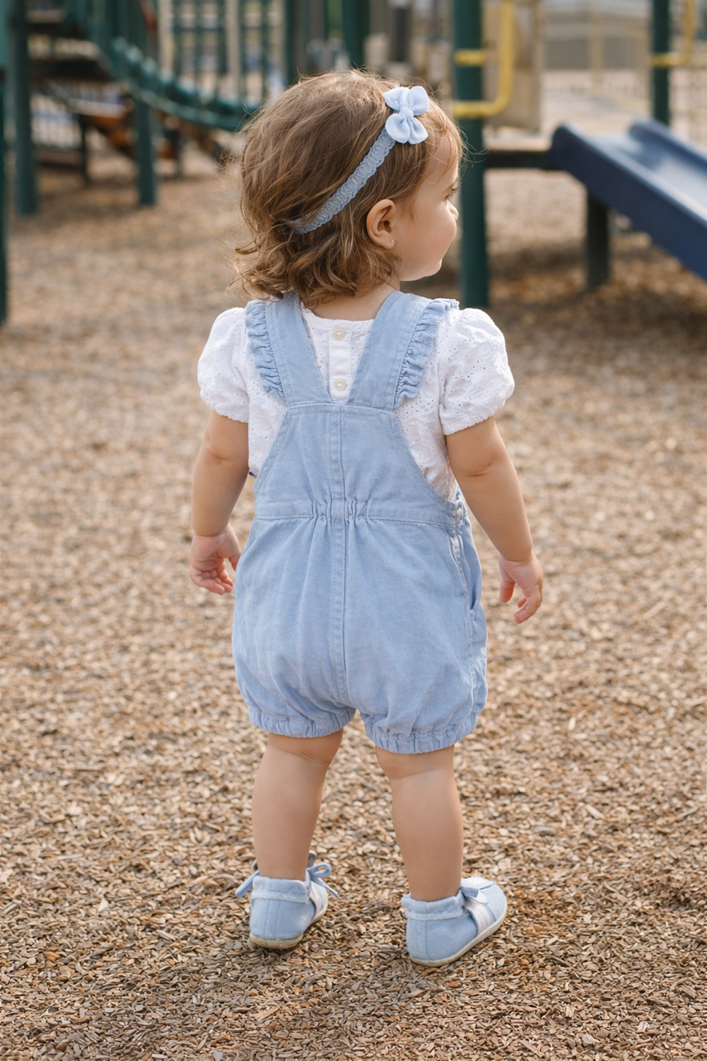 Back view of toddler wearing light blue dungaree and white top, standing at playground, showing elastic waist and straps.