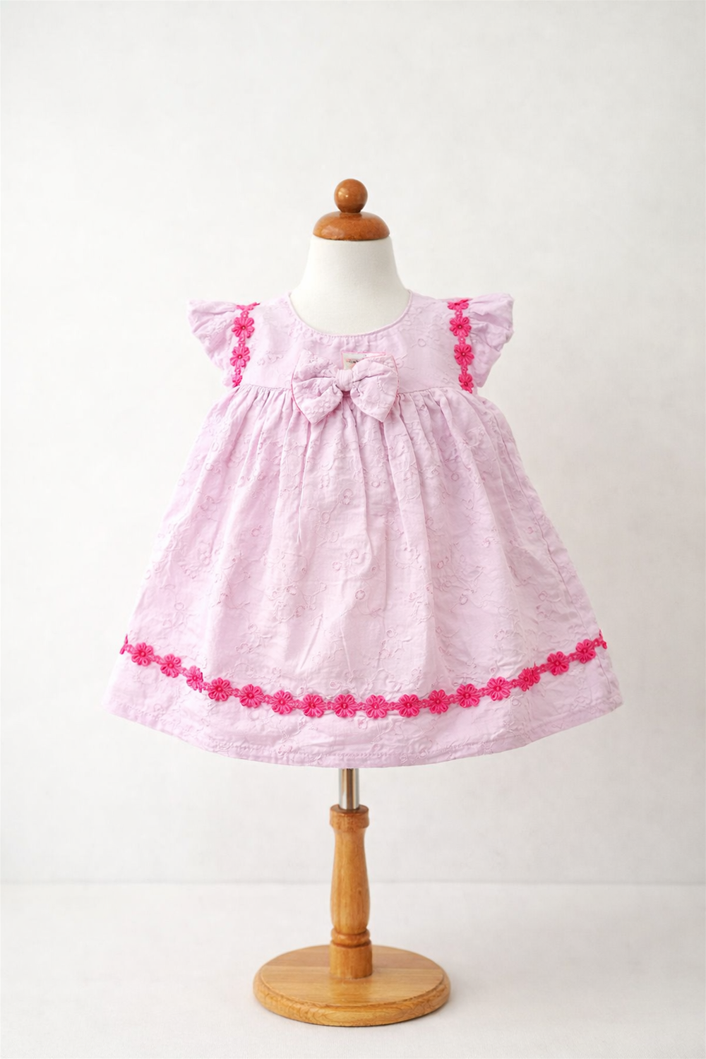lavender baby dress displayed on a mannequin, featuring lace fabric, rose floral trim, bow detail, and sleeveless design.