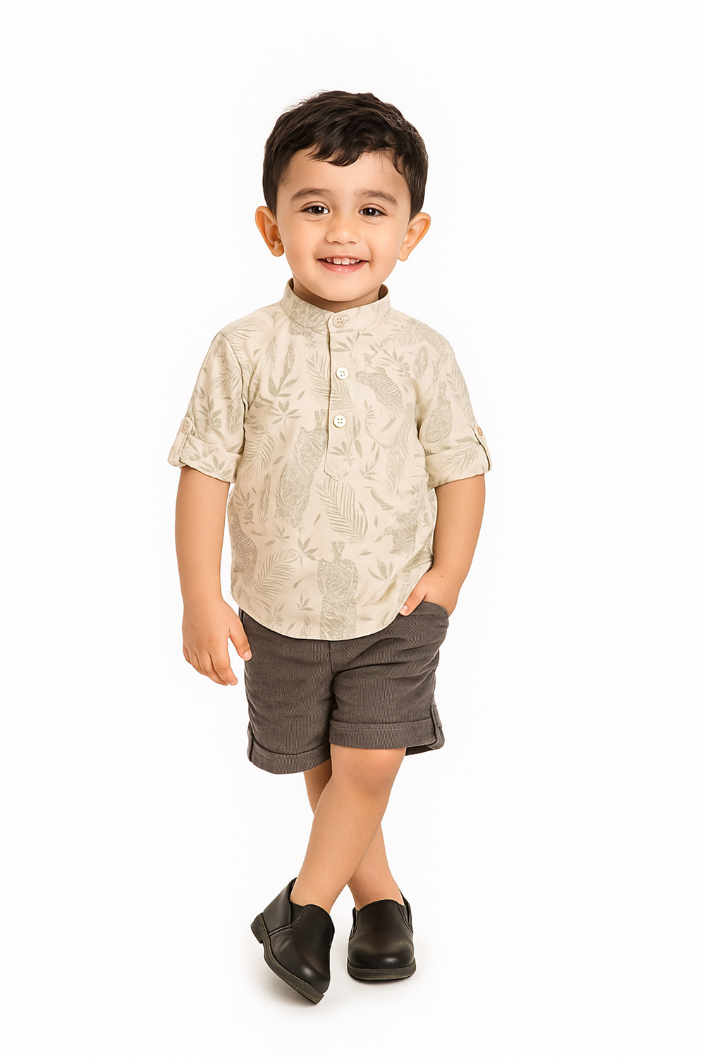 Smiling toddler in beige printed shirt and dark shorts, standing confidently in studio with crossed legs and black shoes.