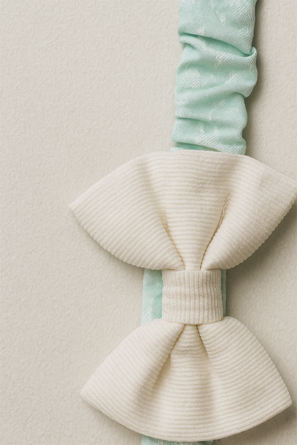 Close-up of cream bow on mint-green elastic hairband, showing fine ribbed fabric texture and soft pastel design background.