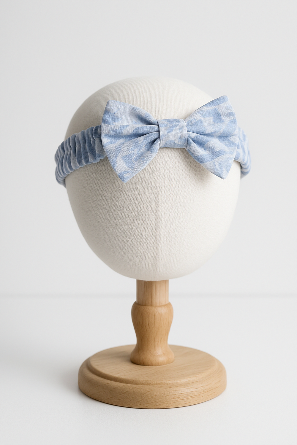 A soft, stretchy baby headband featuring a large bow in a light blue tie-dye pattern. The fabric is textured and ruched for comfort, designed to fit snugly while adding a charming touch to any baby outfit.