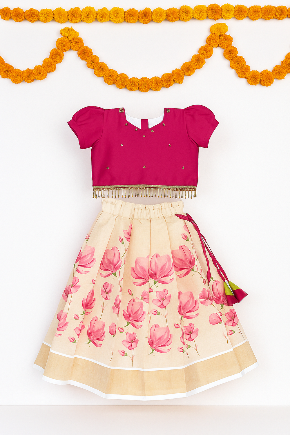 A festive kidswear set with pink blouse and beige skirt featuring bold pink floral prints, displayed against white background with garlands.