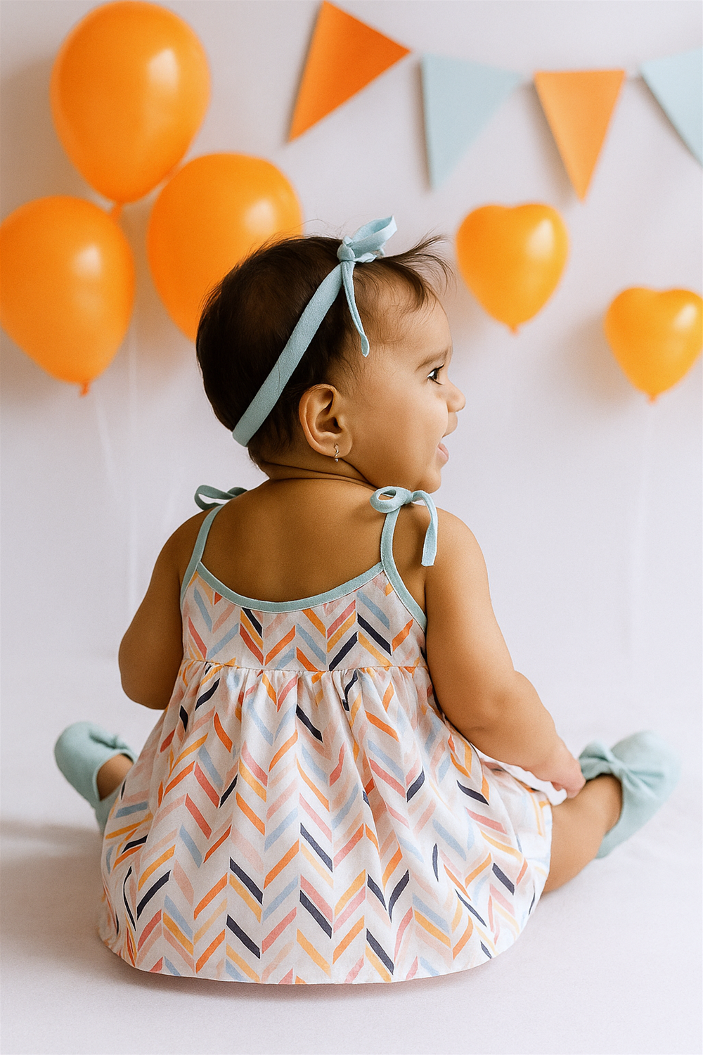 A baby girl sits facing away, showcasing the back of her colorful chevron-print dress with teal trim and tie-up shoulder straps. The festive background features bright orange balloons and pastel bunting, creating a cheerful celebration vibe.