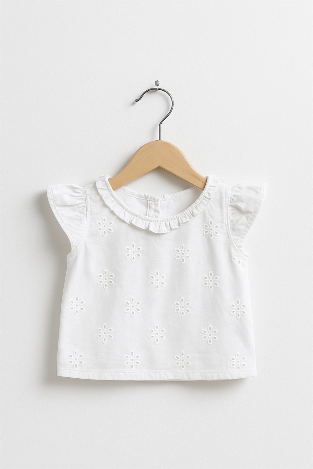 White sleeveless embroidered cotton top on a wooden hanger, featuring flutter sleeves, floral cutwork design, and soft ruffled neckline detailing.