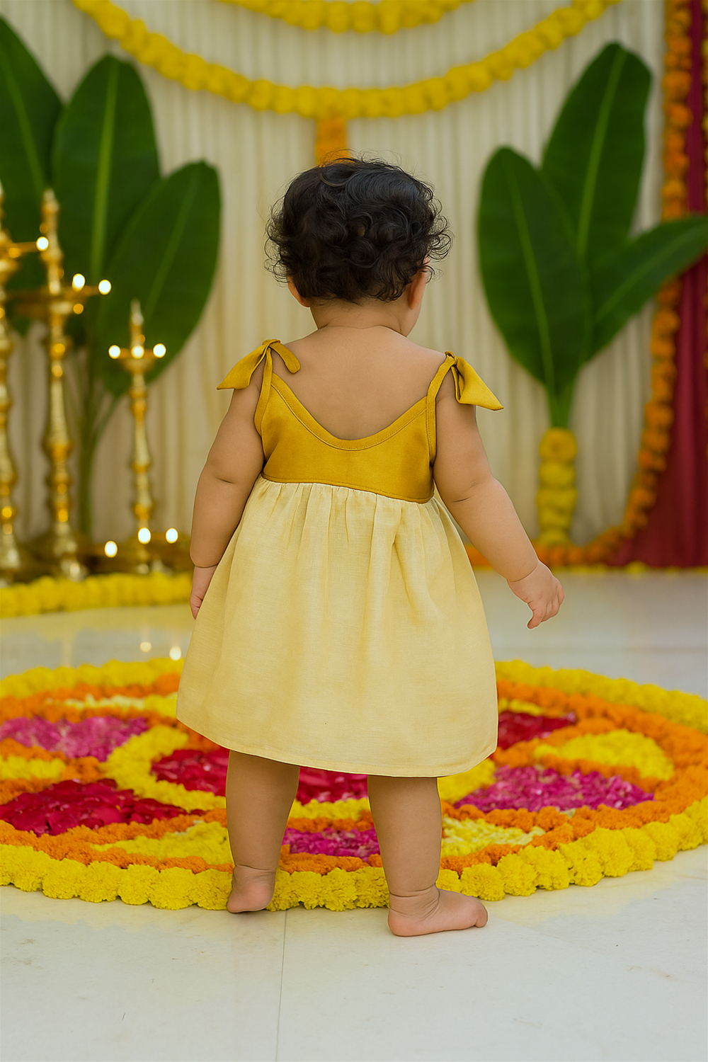 A baby dressed in a mustard yellow and cream Onam frock is seen from the back, standing near a vibrant floral pookalam. The festive backdrop with banana leaves, marigold garlands, and traditional lamps enhances the celebratory mood.