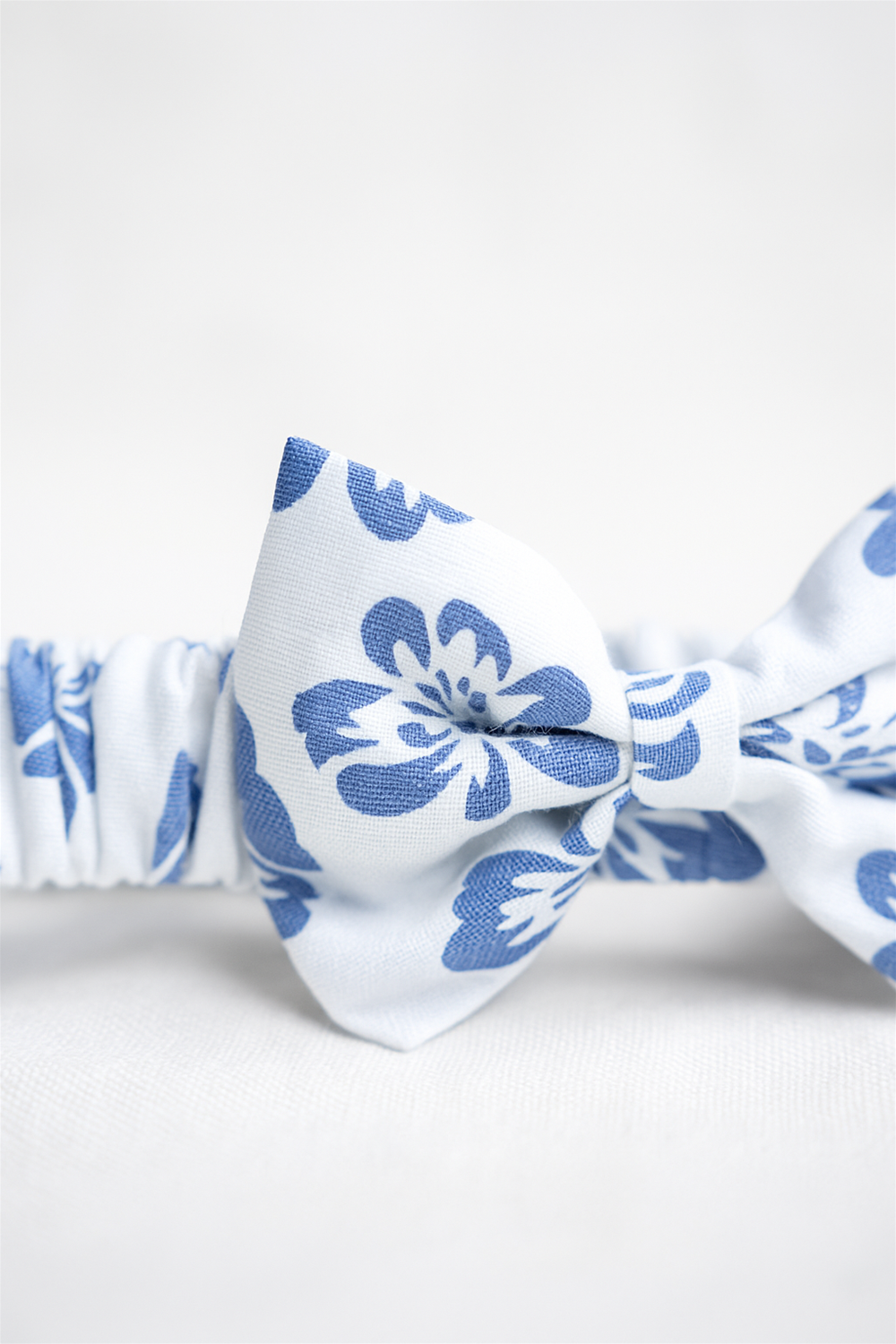 A white elastic headband featuring a navy-blue floral print, designed with a central bow for a stylish and charming look. Perfect as an accessory for babies and toddlers, combining comfort with elegance.