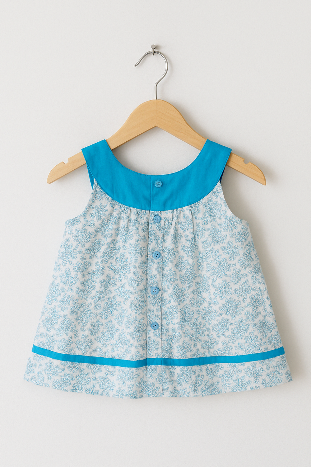 Back view of baby frock with blue buttons and collar, displayed on wooden hanger against white wall background.