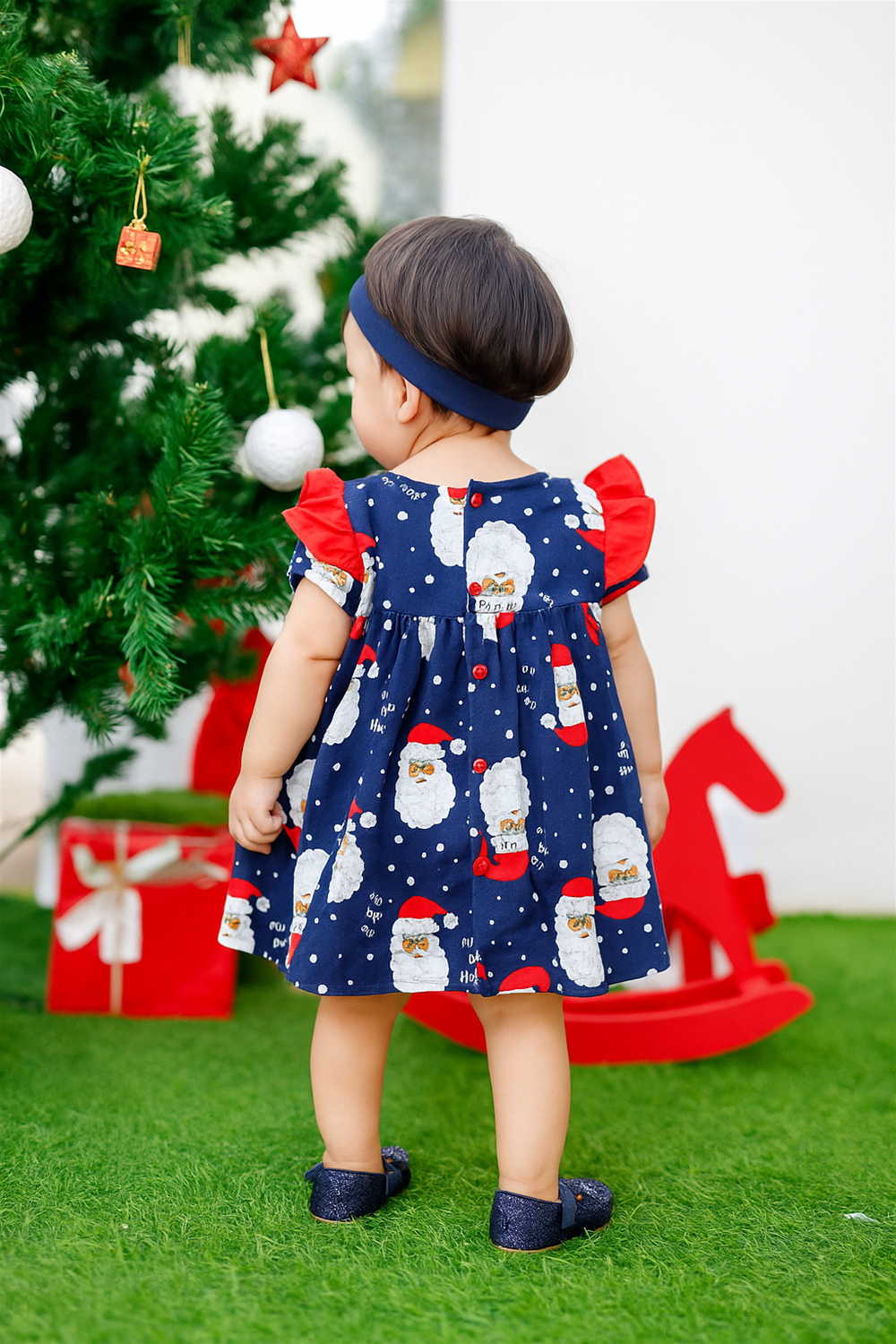 A baby wearing a navy blue Santa print frock with red sleeves, standing near a decorated Christmas tree with gifts.