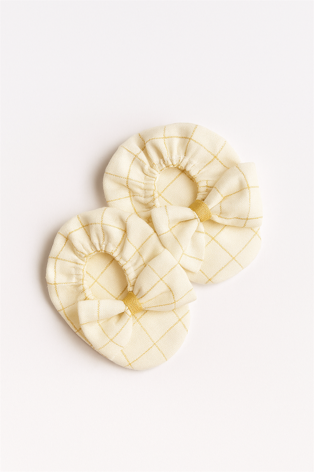 A pair of off-white baby booties with a mustard yellow grid pattern is neatly displayed on a white background. Each bootie is decorated with a soft bow at the front, adding a touch of charm.