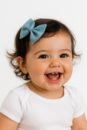 Adorable smiling baby girl wearing aqua blue bow hair clip, curly hair styled naturally, captured in bright studio lighting.