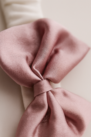 A close-up view of a mauve satin bow attached to a white elastic hairband shows the smooth, glossy texture of the fabric. The soft folds and sheen highlight its refined and delicate craftsmanship.