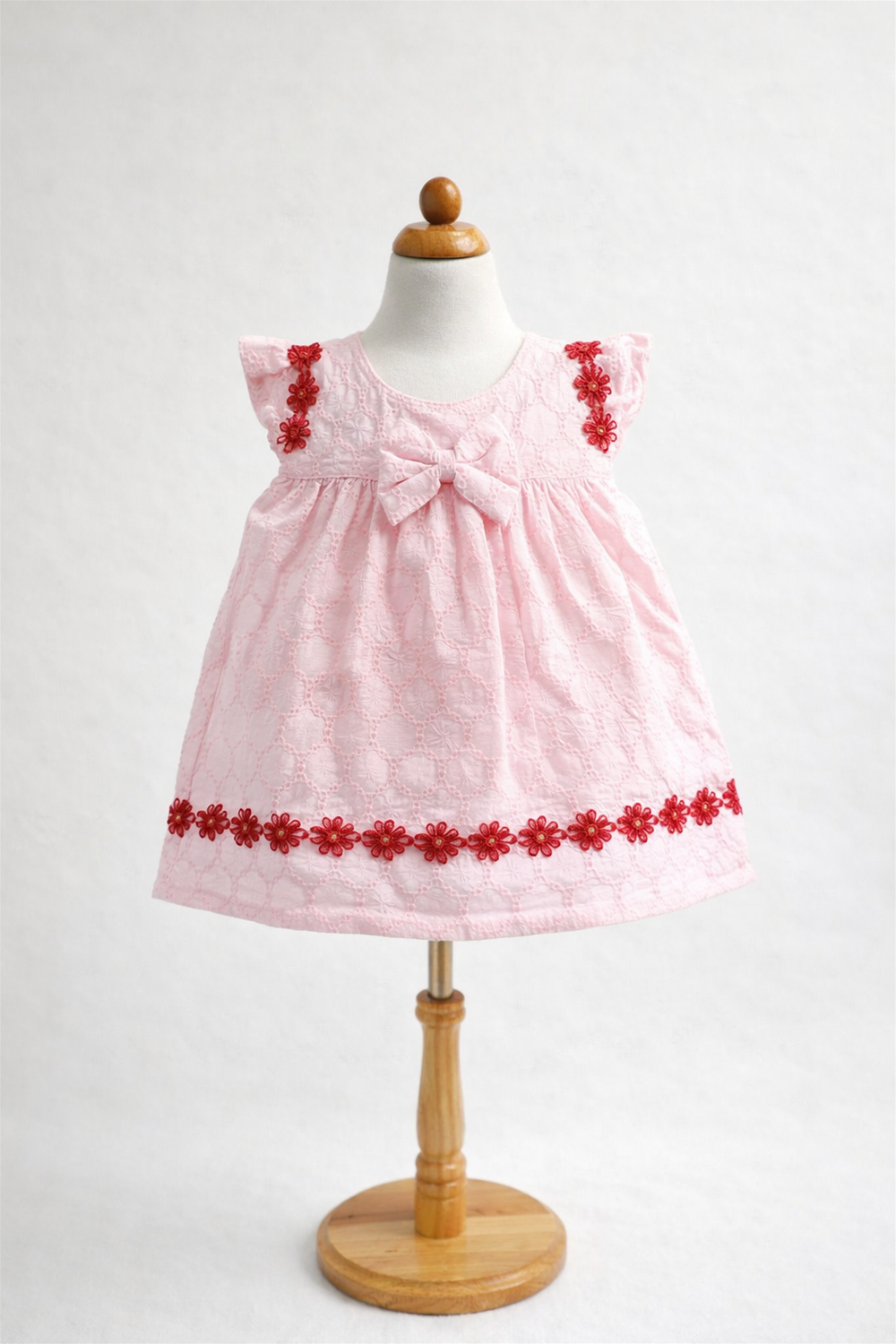 Pink baby dress displayed on a mannequin, featuring lace fabric, red floral trim, bow detail, and sleeveless design