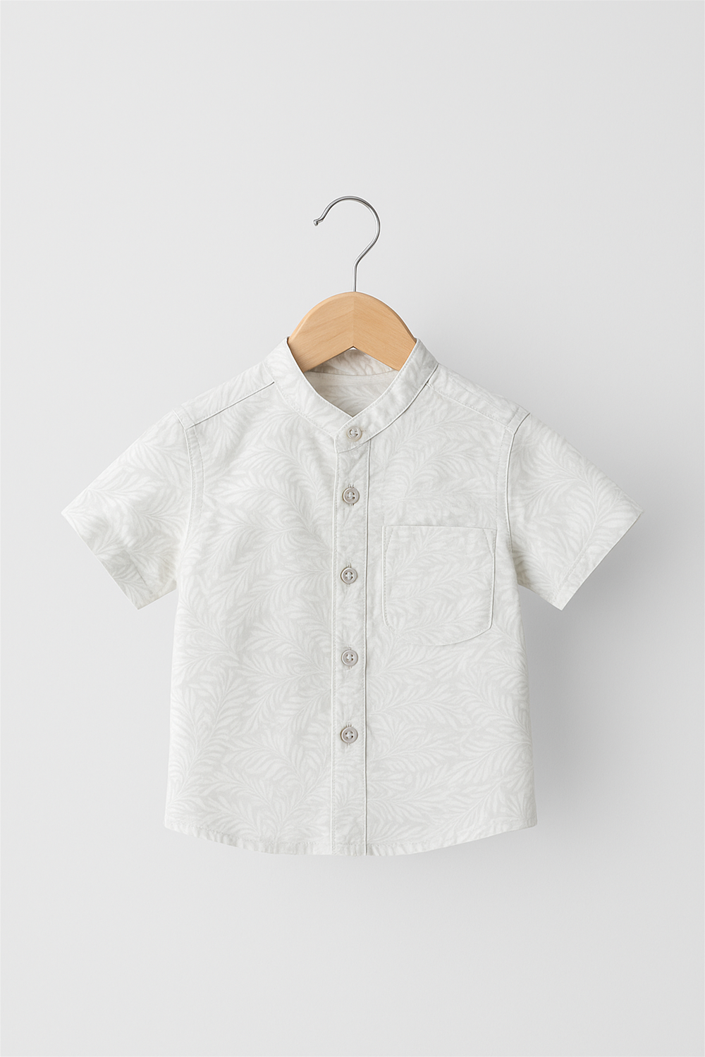 A neatly displayed half-sleeve Chinese collar shirt for boys in soft white, featuring a subtle leafy all-over print. The shirt is buttoned down the front with a chest pocket, hung on a wooden hanger against a plain studio background.
