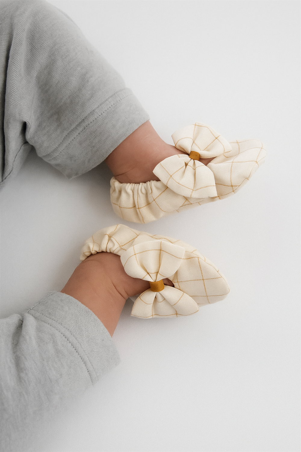 A baby is wearing the same off-white booties with a mustard yellow checkered design and a bow on top. The shoes fit snugly with elastic around the ankles, ensuring comfort and security.