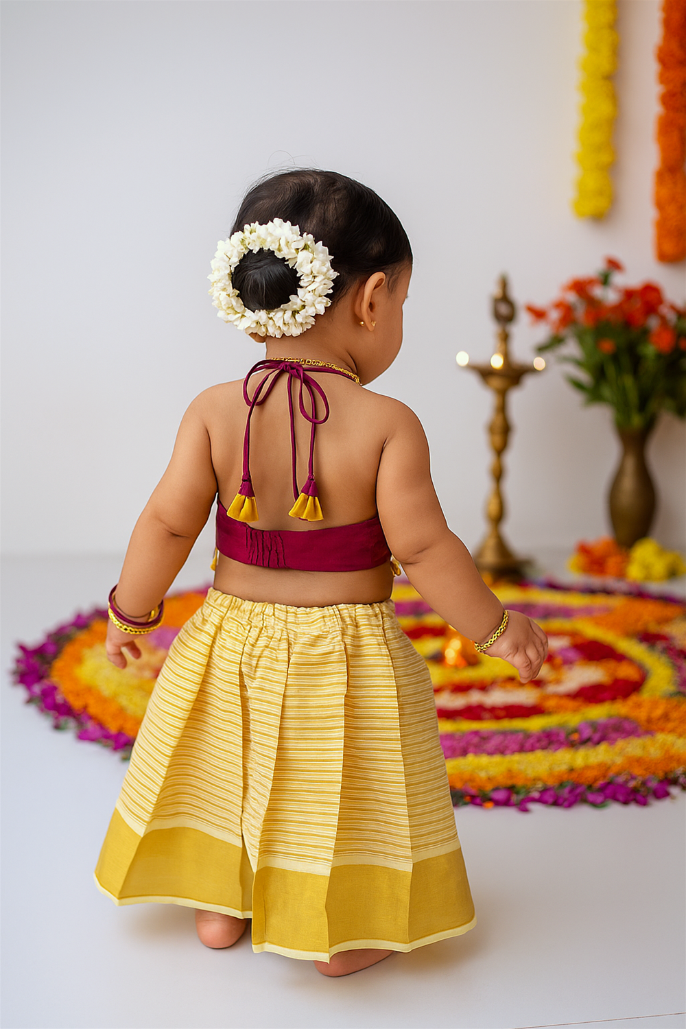 The baby girl is seen from the back, her maroon halter top tied with tassels and her hair adorned with jasmine garlands. She stands near a colorful flower rangoli and traditional brass lamp with marigold garlands in the background.