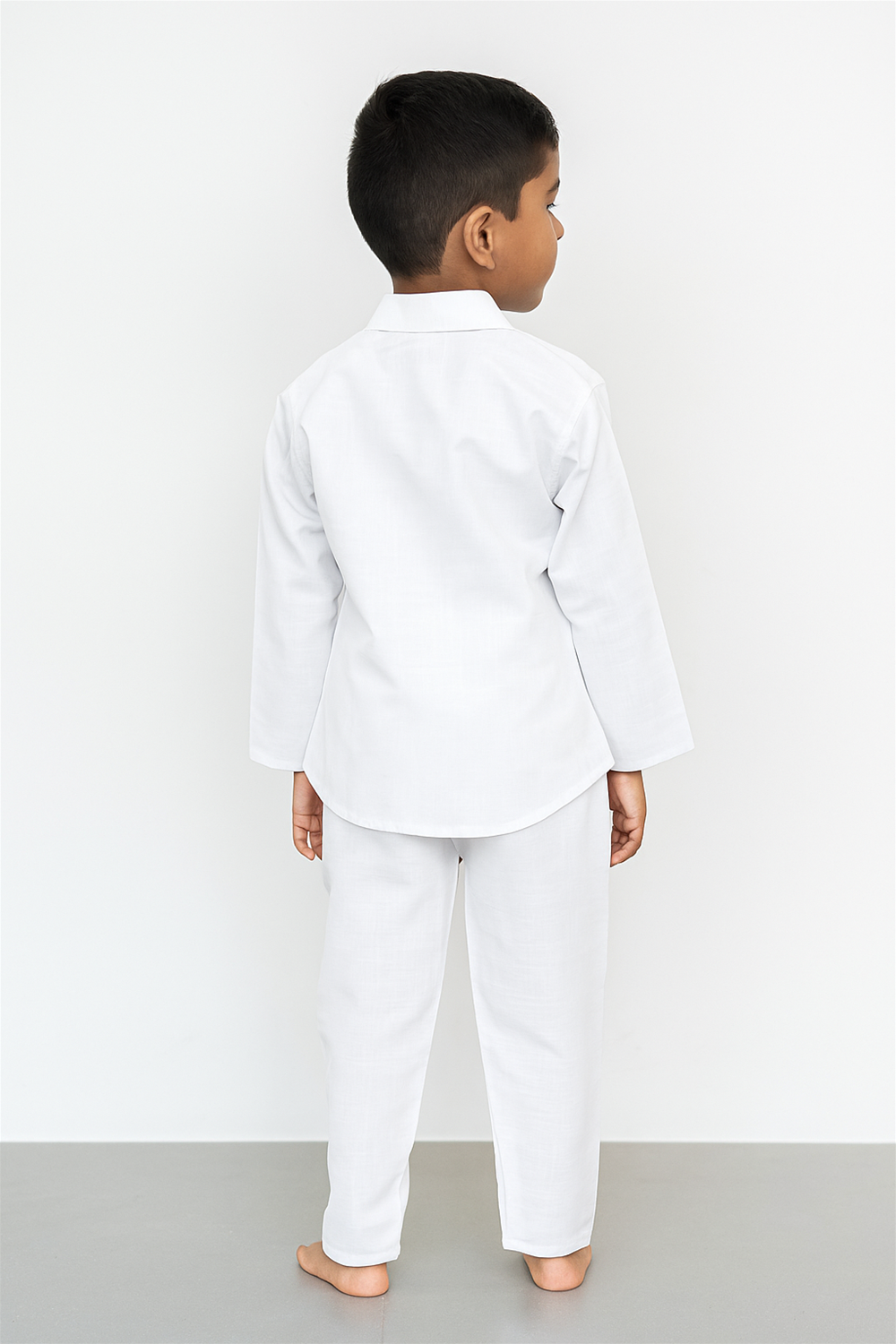 A young boy is standing barefoot, facing away from the camera, wearing a plain white long-sleeved kurta and matching white trousers. The outfit is simple and traditional, with a clean, minimal design against a plain light background.
