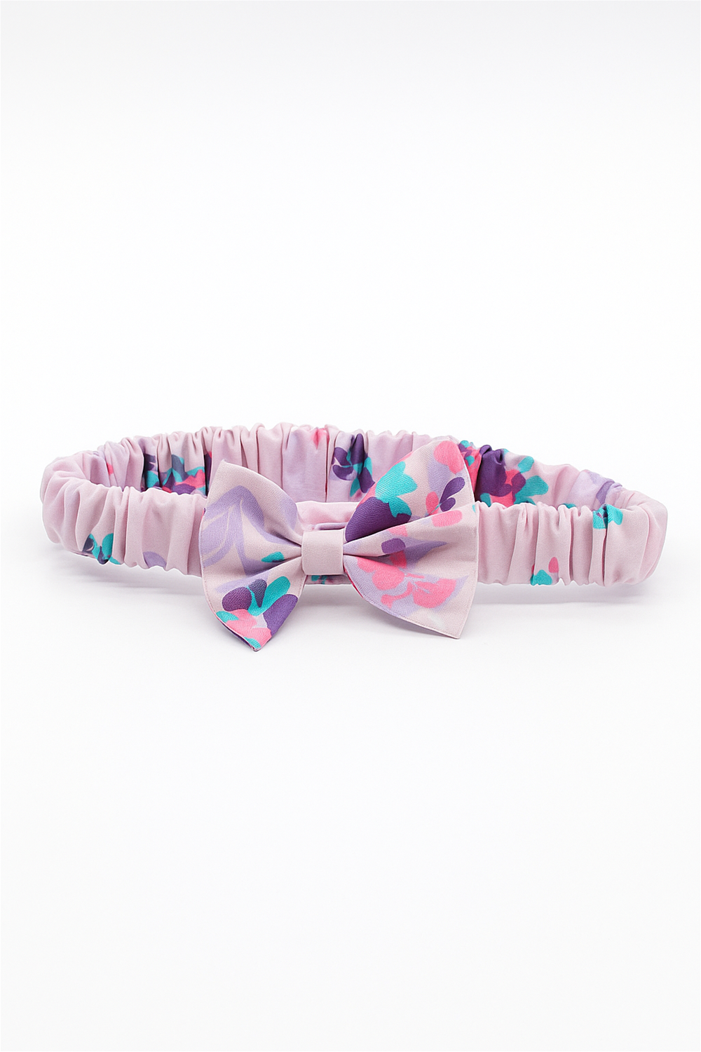 A pink elastic baby headband with a gathered fabric band, featuring a decorative bow in a pastel floral pattern with shades of purple, turquoise, and pink. The headband is laid flat against a white background, showcasing its design in full detail.