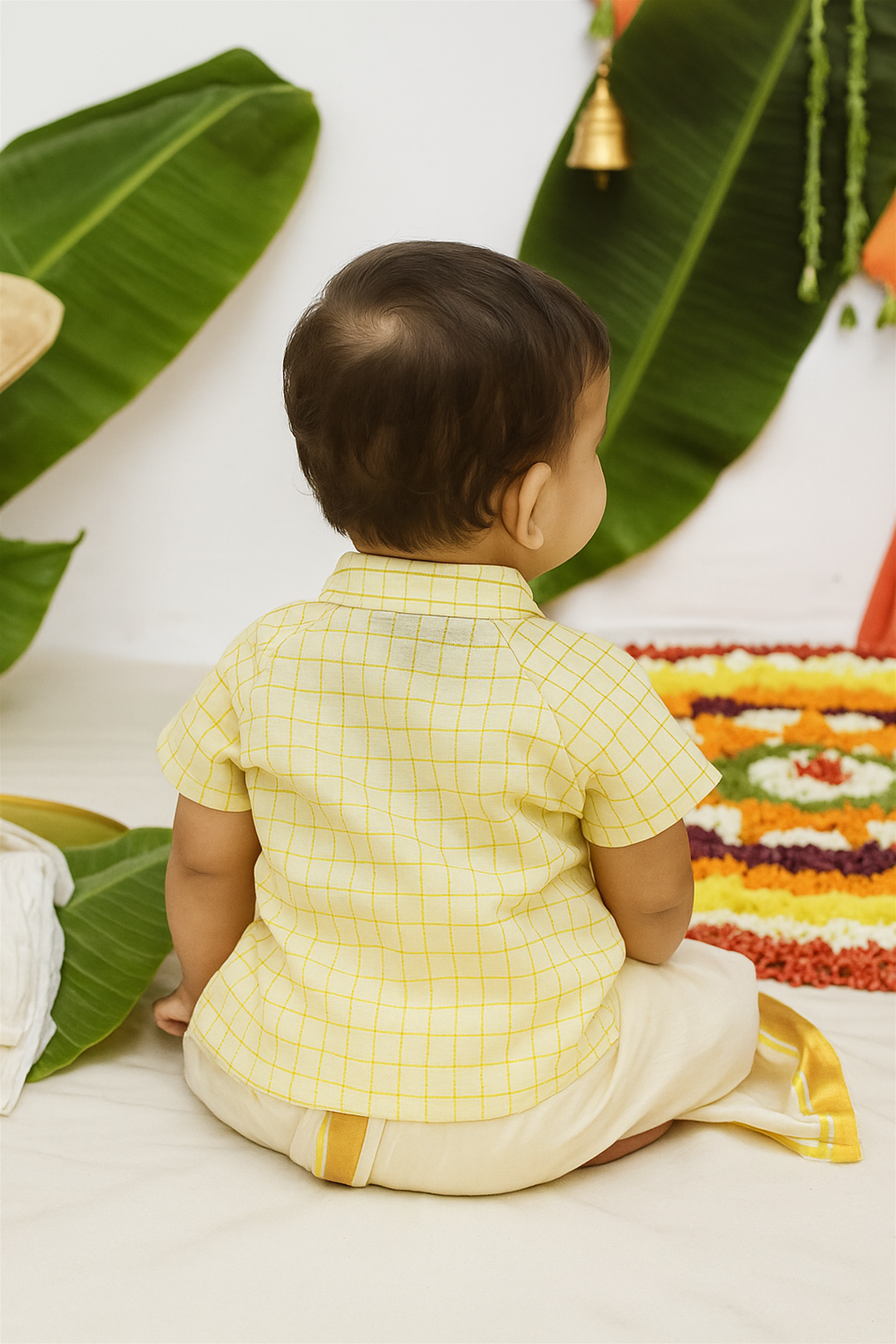 Baby boy from back wearing yellow checked shirt and cream dhoti, sitting before colorful floral rangoli and green banana leaves.