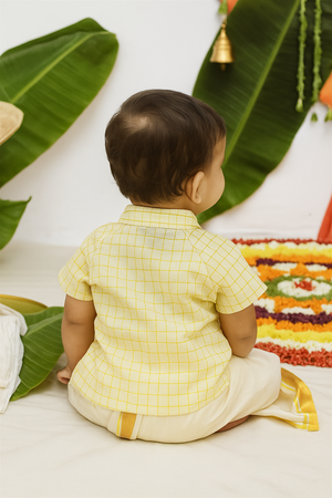 Baby boy from back wearing yellow checked shirt and cream dhoti, sitting before colorful floral rangoli and green banana leaves.
