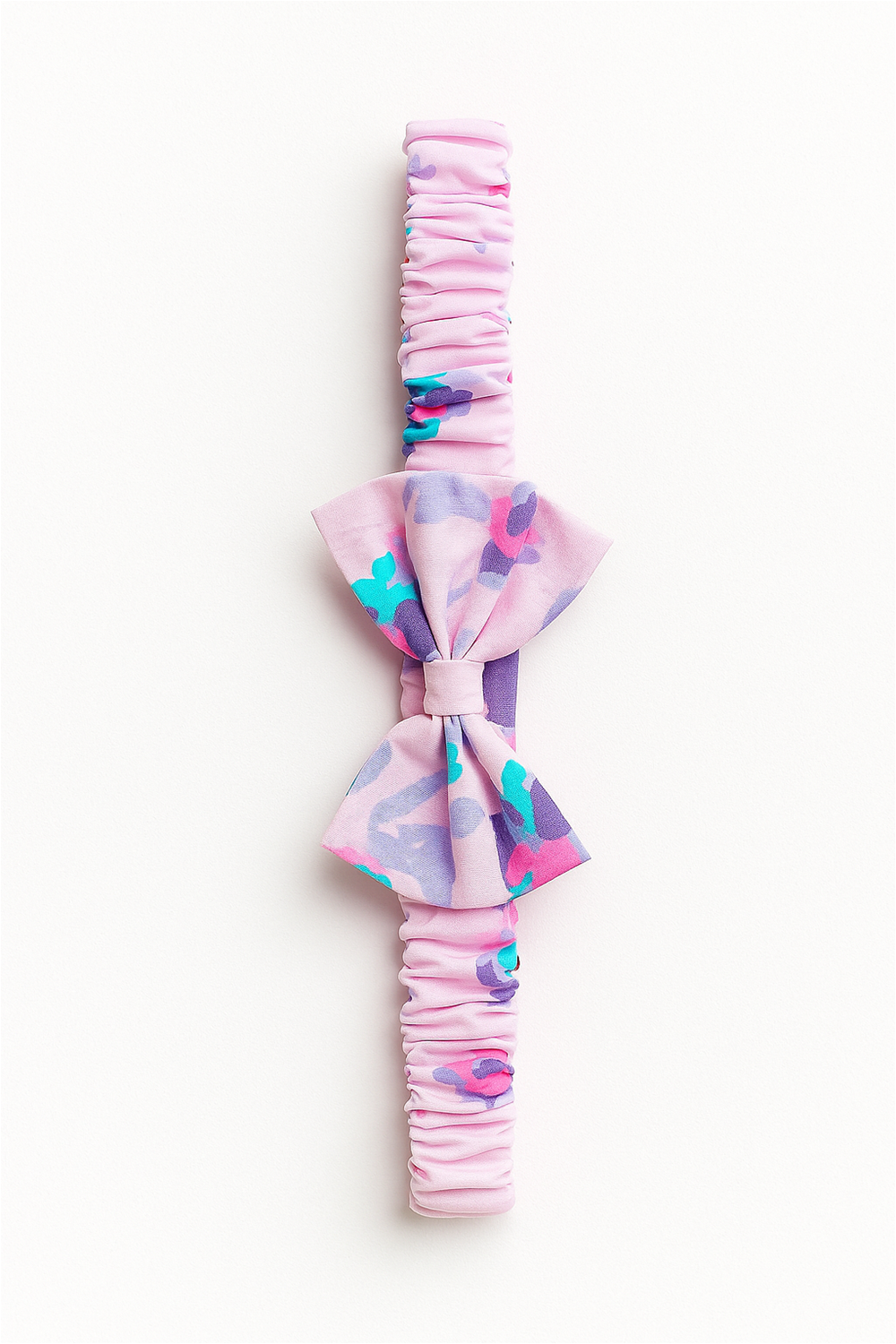 A pink elastic baby headband with a gathered fabric band, featuring a decorative bow in a pastel floral pattern with shades of purple, turquoise, and pink. The headband is laid flat against a white background, showcasing its design in full detail.