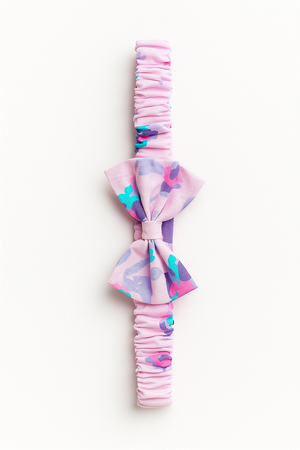 A pink elastic baby headband with a gathered fabric band, featuring a decorative bow in a pastel floral pattern with shades of purple, turquoise, and pink. The headband is laid flat against a white background, showcasing its design in full detail.
