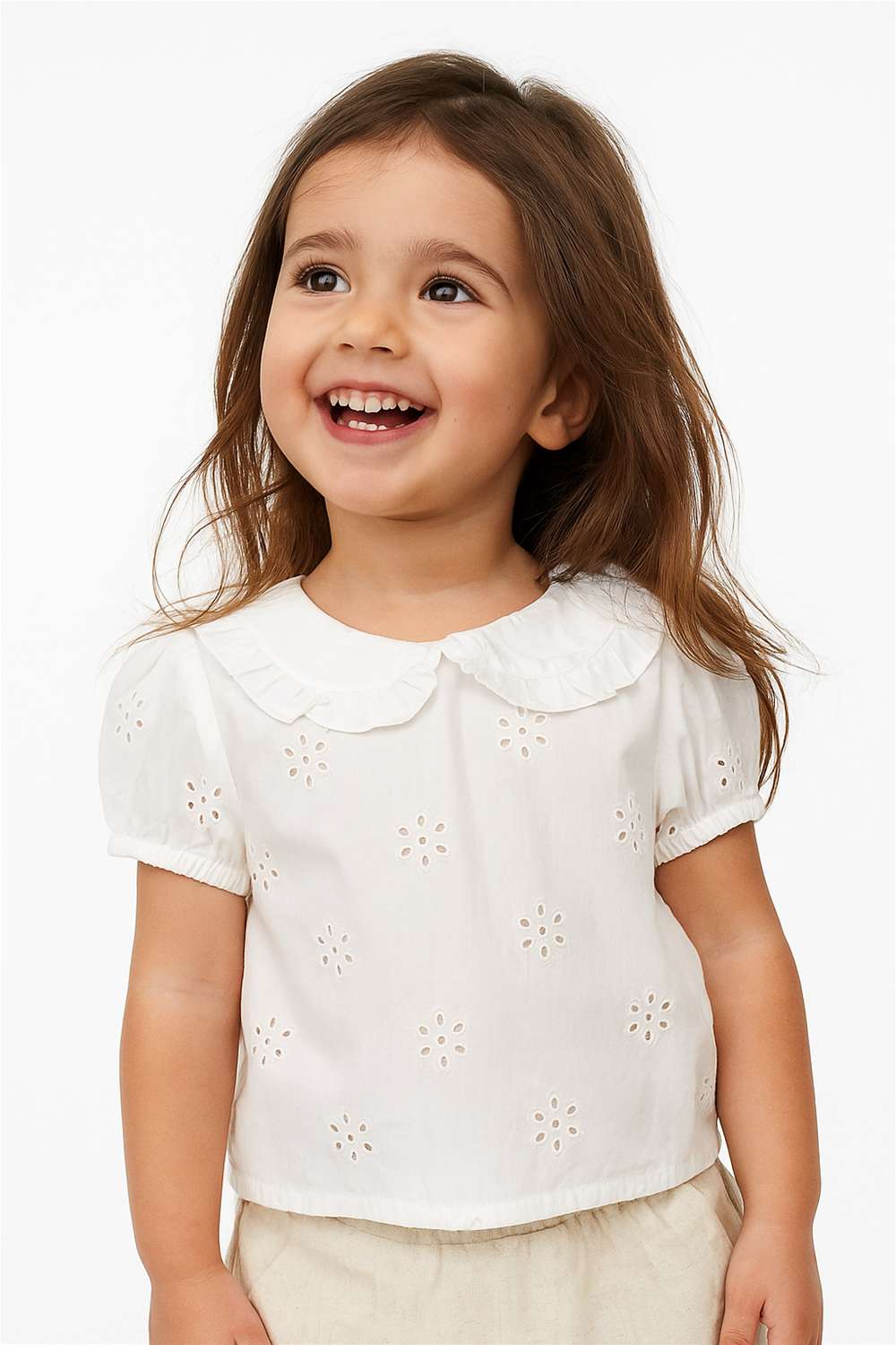 A young girl with long brown hair smiles brightly while looking slightly upward. She is wearing a white short-sleeved top with eyelet flower patterns and a ruffled collar, paired with light beige bottoms.