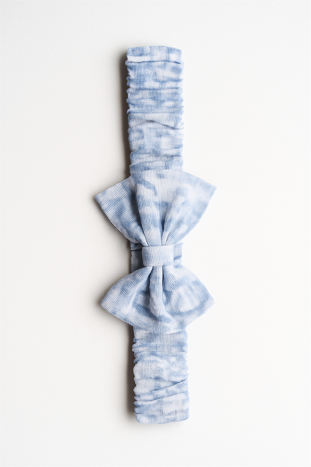 A soft, stretchy baby headband featuring a large bow in a light blue tie-dye pattern. The fabric is textured and ruched for comfort, designed to fit snugly while adding a charming touch to any baby outfit.