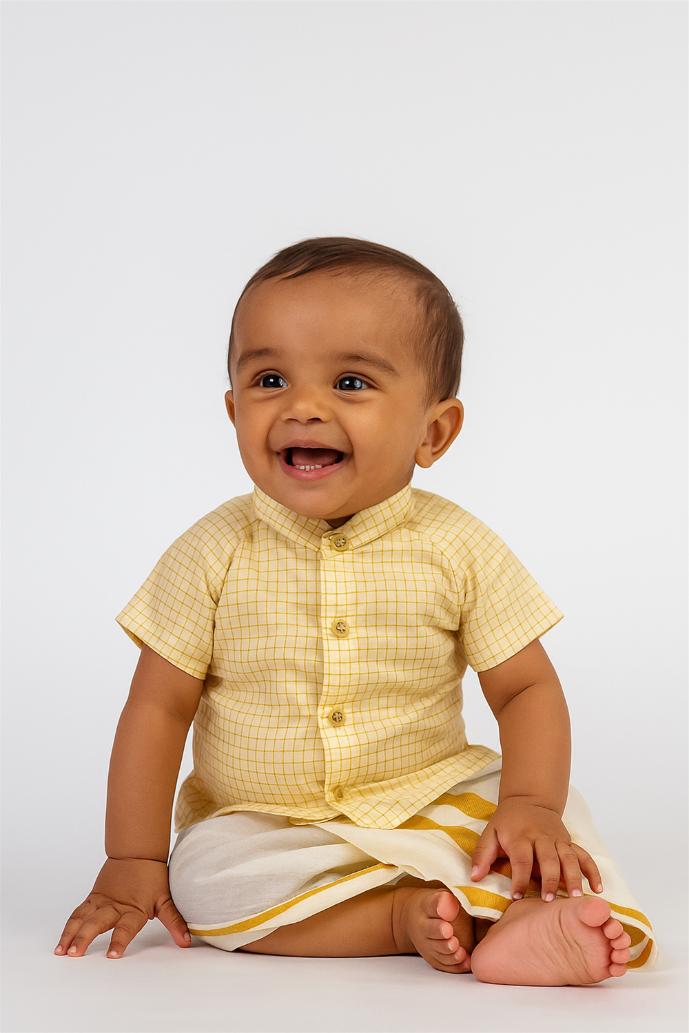 Cute baby boy in yellow checkered shirt and traditional white-gold bordered dhoti, smiling joyfully against a studio background.