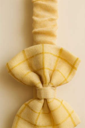 A detailed close-up of a yellow checkered fabric bow on an elastic hairband. The texture and fine weave of the material are clearly visible, emphasizing its soft handcrafted look.