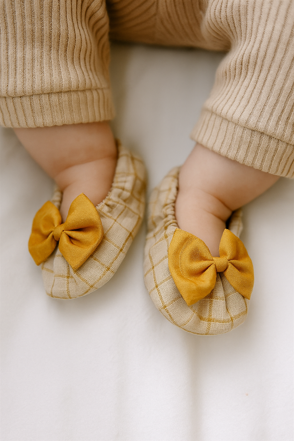 A baby wears beige checkered booties with bright golden bows, complementing the cozy ribbed beige outfit. The shoes look lightweight and soft, perfect for keeping tiny feet warm and stylish.