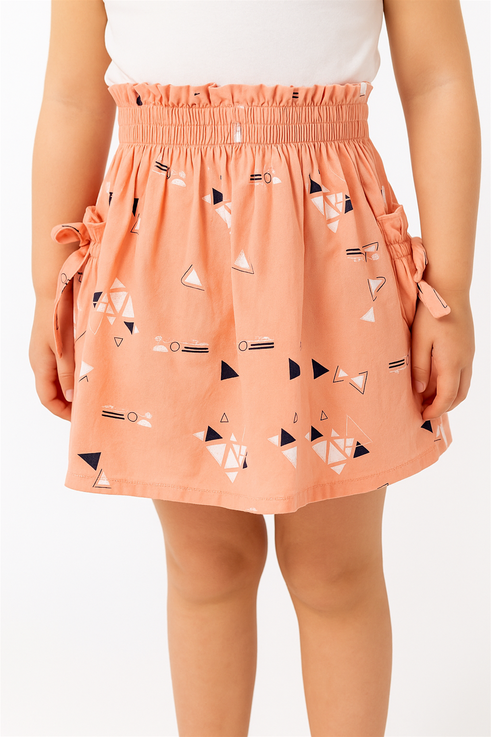 Close-up of a peach-colored skirt with geometric prints, elastic waistband, and side bows, styled on a child model.