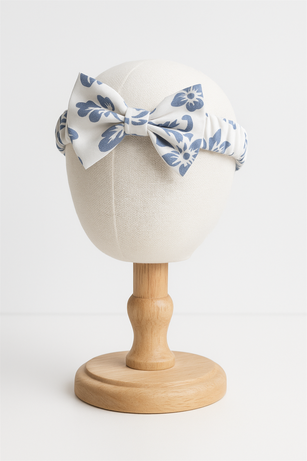 A white elastic headband featuring a navy-blue floral print, designed with a central bow for a stylish and charming look. Perfect as an accessory for babies and toddlers, combining comfort with elegance. In head foam mannequin.