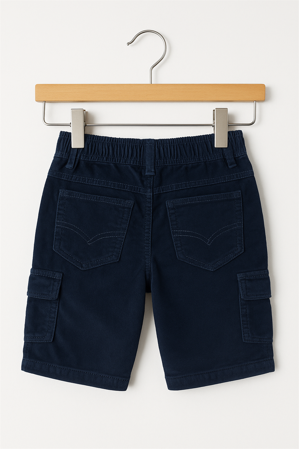 A pair of navy blue corduroy cargo shorts for children displayed on a wooden hanger. The shorts feature a metal button closure, belt loops, front and back pockets, and side flap cargo pockets with metallic snap buttons. The background is a plain off-white.