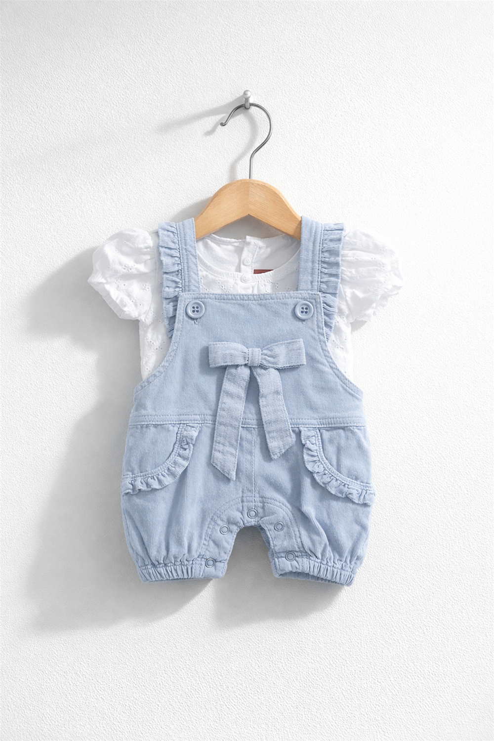 Front view of light blue baby dungaree featuring bow detail, ruffled pockets, snap buttons, styled with white puff sleeve top.