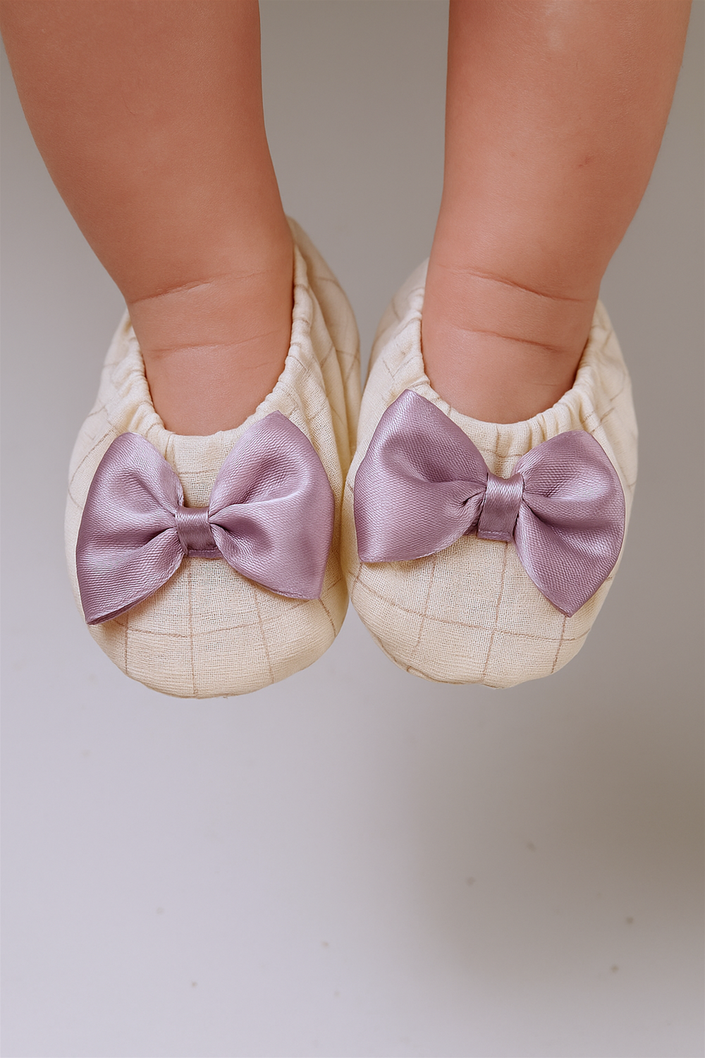 A baby is wearing the cream checkered booties with lavender bows, their tiny feet visible inside the soft shoes. The bows stand out with a gentle sheen, adding a charming touch.