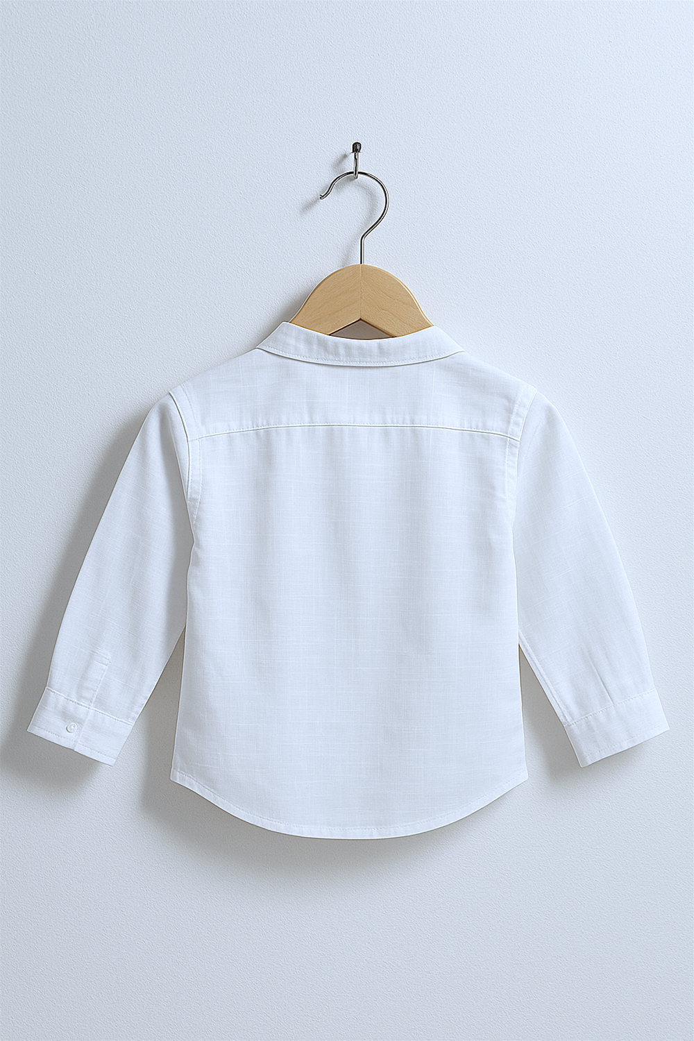 Alternative text: Back view of a long-sleeved white shirt for children, displayed on a wooden hanger against a light wall. The shirt features a straight back yoke and buttoned cuffs, photographed in bright studio lighting.