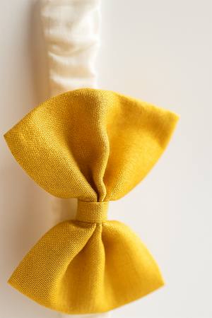 A close-up image of a textured golden-yellow fabric bow attached to a white elastic band. The shot highlights the woven detail and rich color of the bow, giving it a refined and elegant appearance.