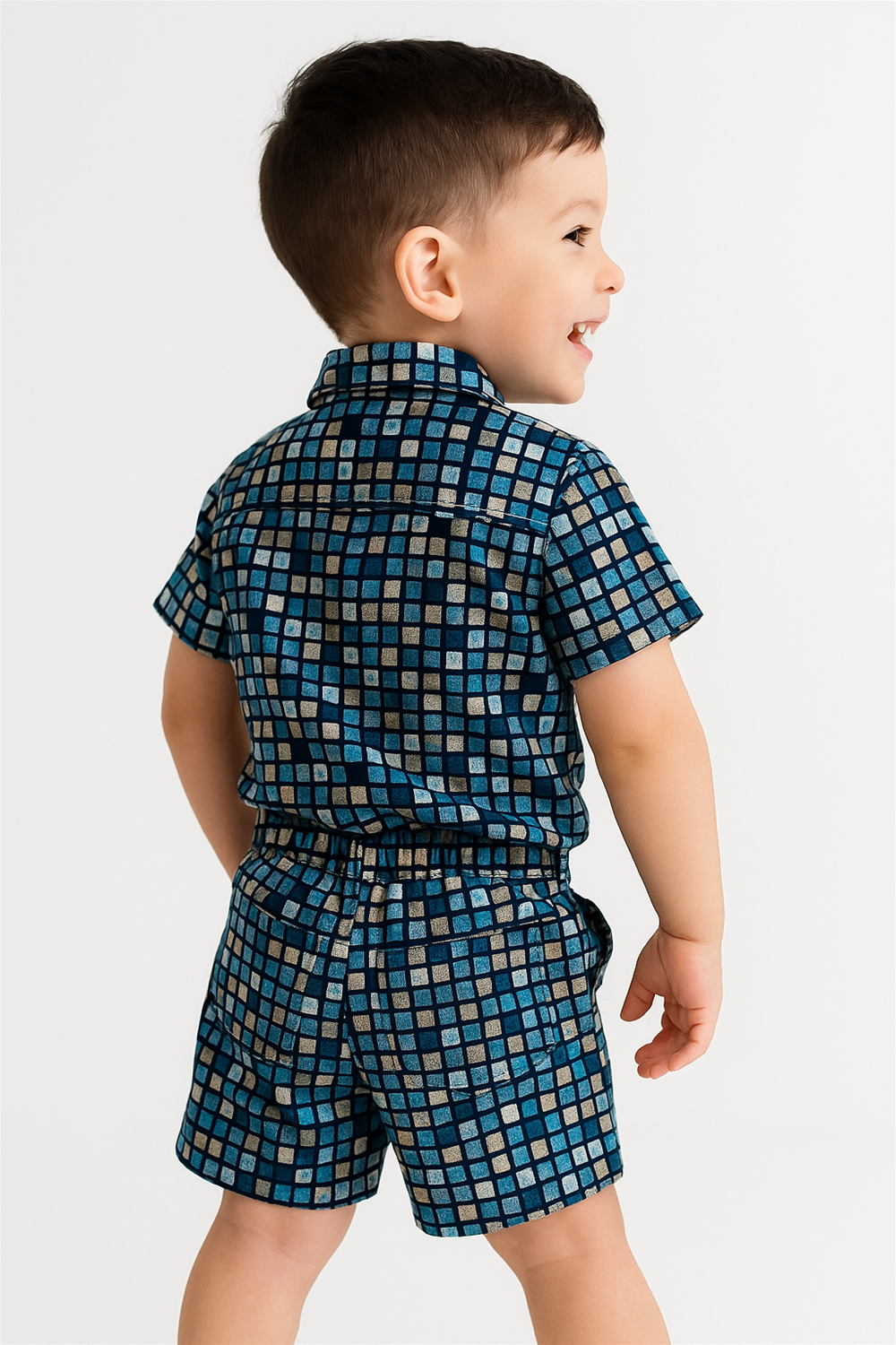 Smiling boy in blue checkered co-ord set, short sleeves and shorts, viewed from back, standing indoors with bright light.